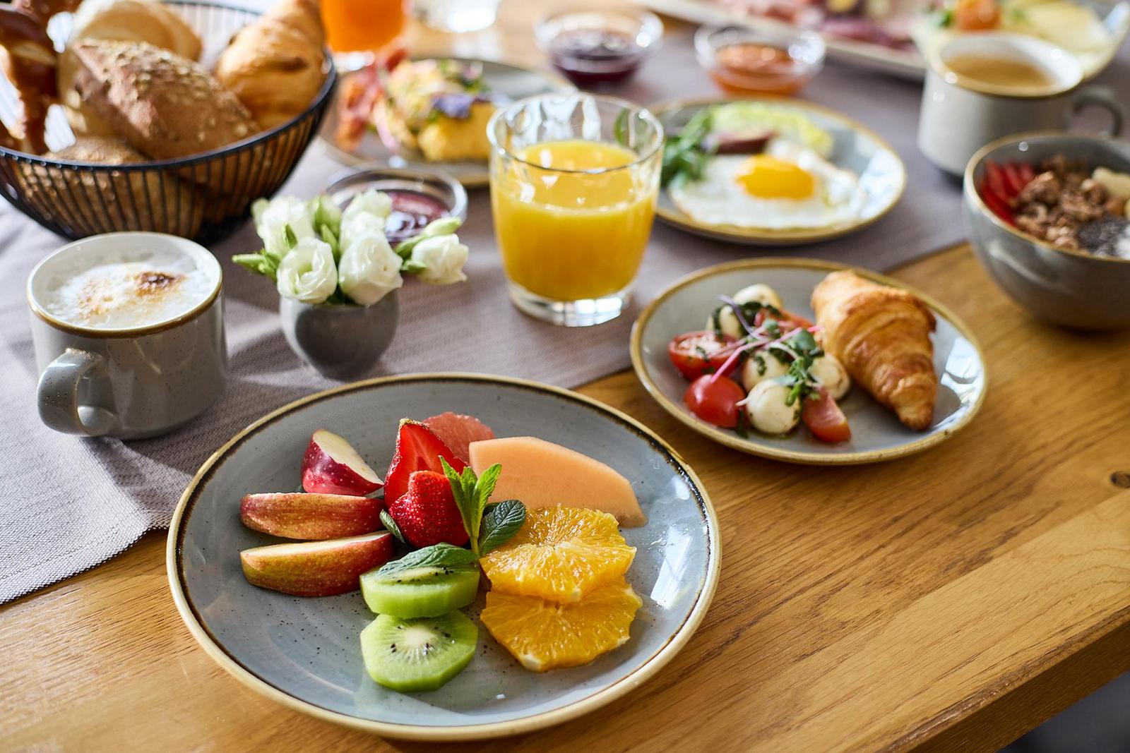 Frühstück im Hotel Krone: Obstteller mit Kiwi, Orange, Erdbeere und Melone, daneben Croissant mit Caprese, plus Orangensaft und Cappuccino auf Holztisch.