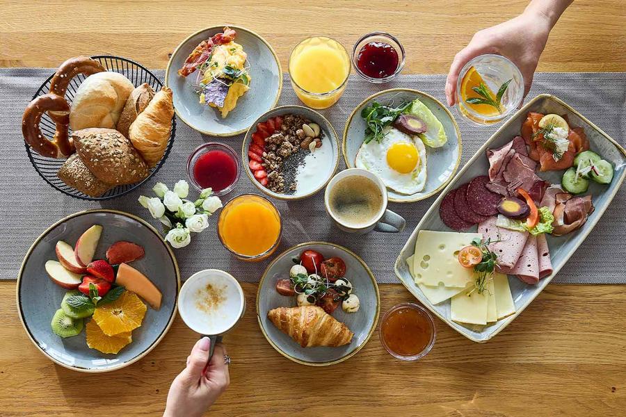 Blick auf einen üppig gedeckten Frühstückstisch. Zwei Personen greifen nach Getränken