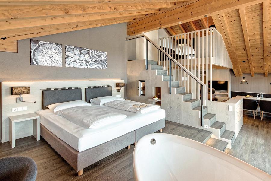 Helle Suite im Hotel Krone: Doppelbett, freistehende Badewanne, Treppe zur Galerie mit Wohnbereich unter sichtbarer Holzbalkendecke.