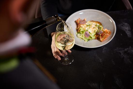 Ein Kellner schenkt ein frisches Glas Weißwein ein. Im Hintergrund ist ein Teller mit Essen zu sehen